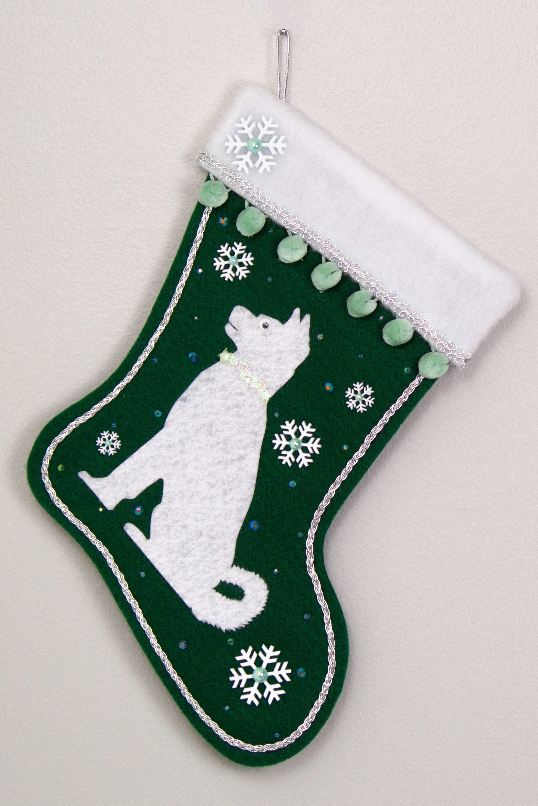 Akita dog in white felt Christmas stocking on  dark green felt with snowflakes, minty green pom poms and decorations in silver and mint green. (Front of stocking)