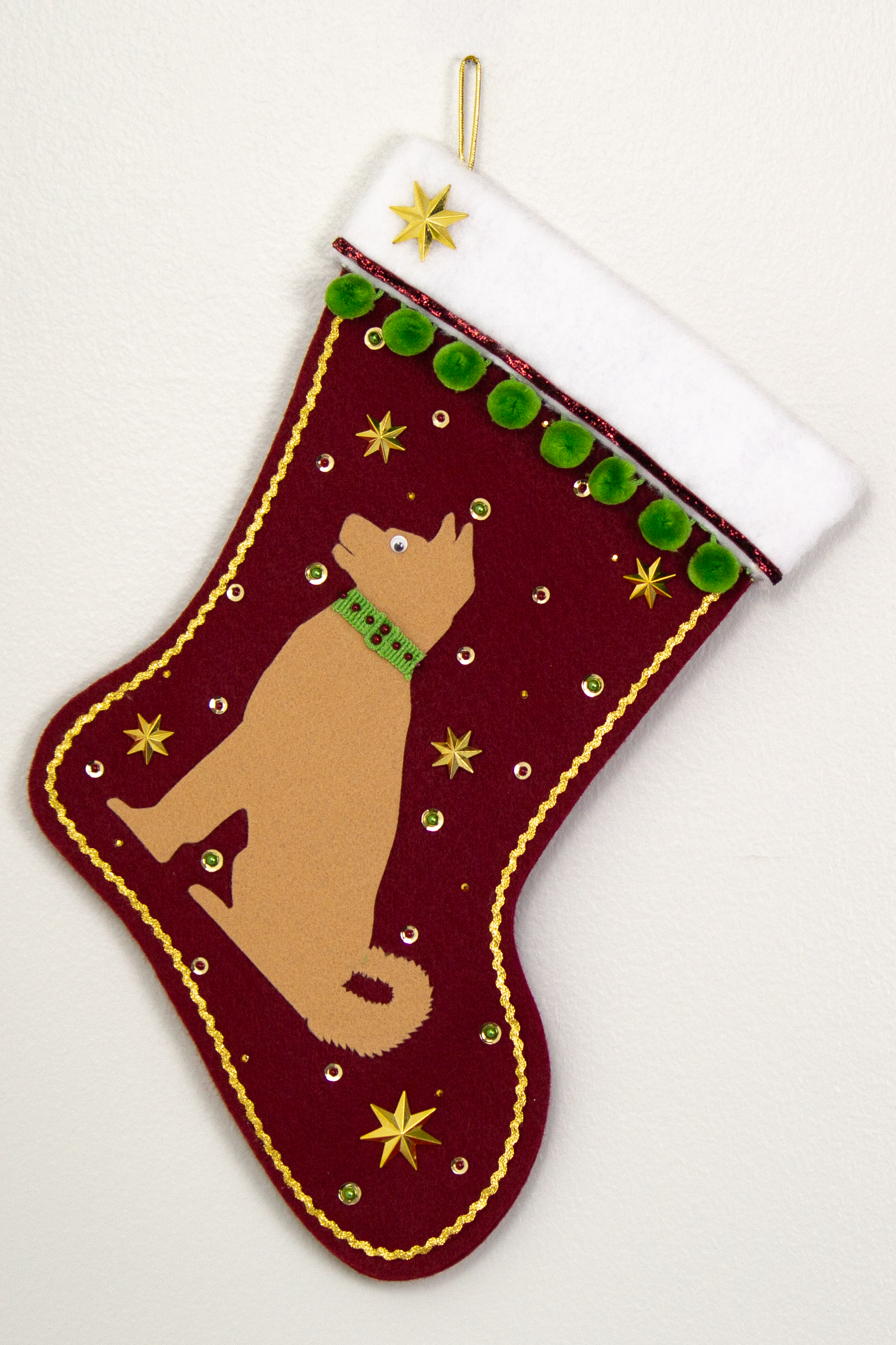 Akita dog in tan felt Christmas stocking on ruby red felt with gold stars, green pom poms and decorations in red, green and gold. (Front of stocking)
