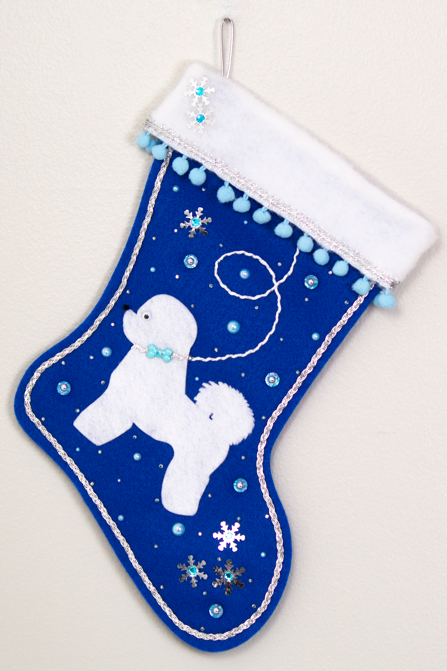 Bichon Frise dog in white felt Christmas stocking on royal blue felt with sequin stars, pom poms and decorations in light blue, white and silver. (Front of stocking)