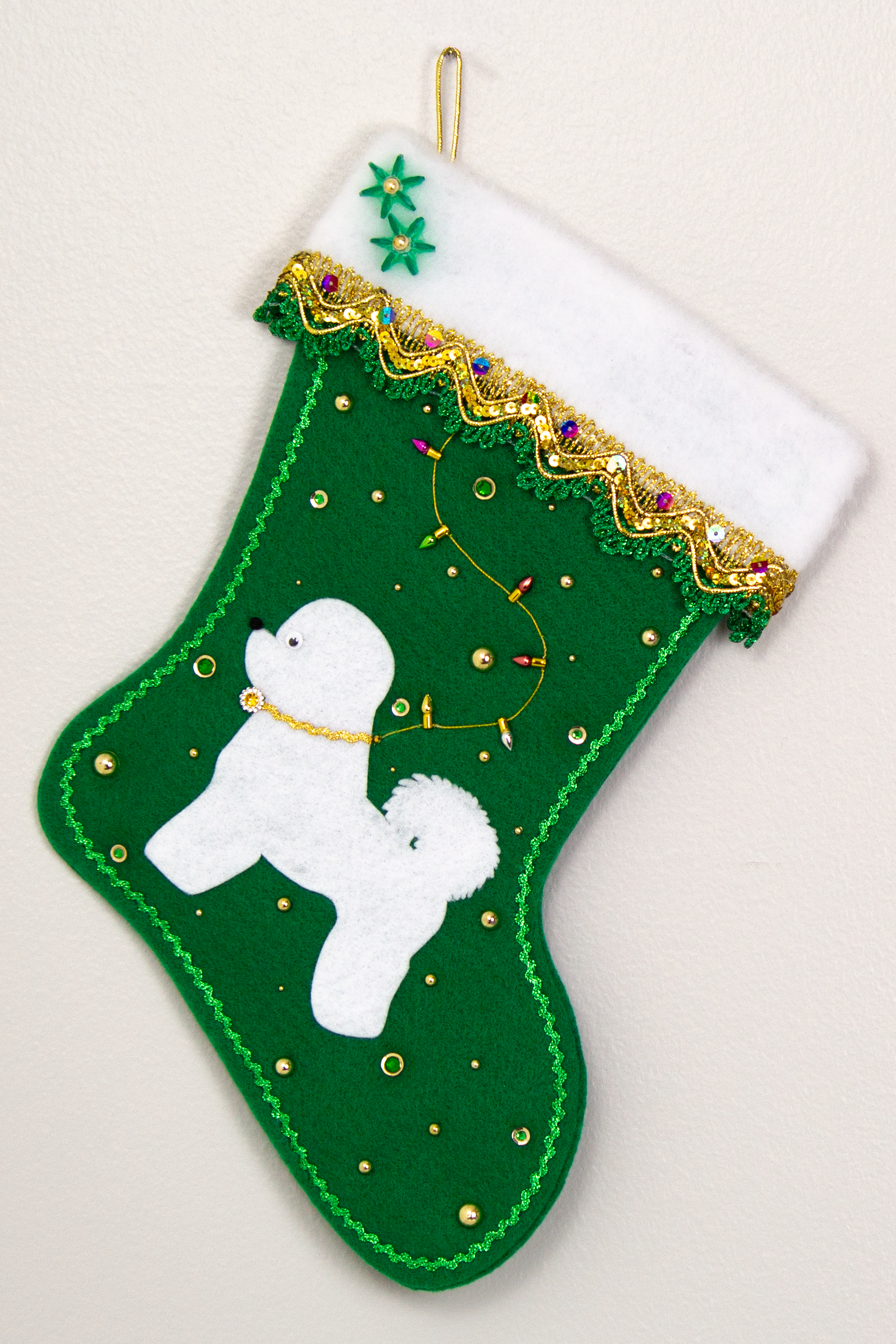 Bichon Frise dog in white felt Christmas stocking on holiday green felt with fancy gold trim, a string of "lights" and decorations in green and gold. (Front of stocking)