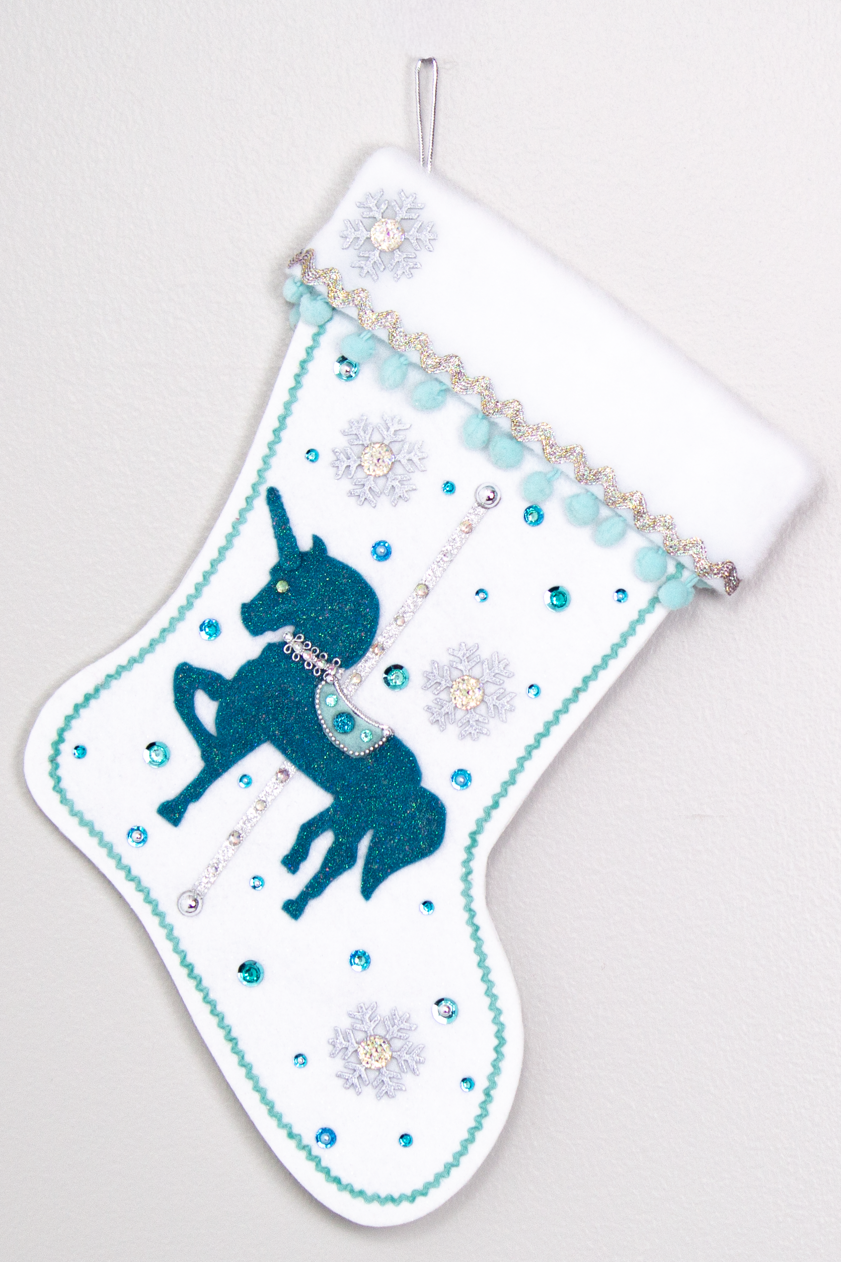 Turquoise felt glittered unicorn on a white glitter felt handmade Christmas stocking with pom poms, snowflake buttons and decorations in aqua, turquoise, white & silver. (front of Carousel Unicorn stocking)