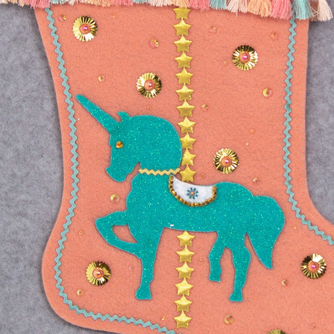 Aqua glittered unicorn on coral felt handmade Christmas stocking with tassel fringe, stars, sunburst sequins and decorated in gold, coral, aqua & multicolored pastels. (front of Carousel Unicorn stocking - close up detail of unicorn)
