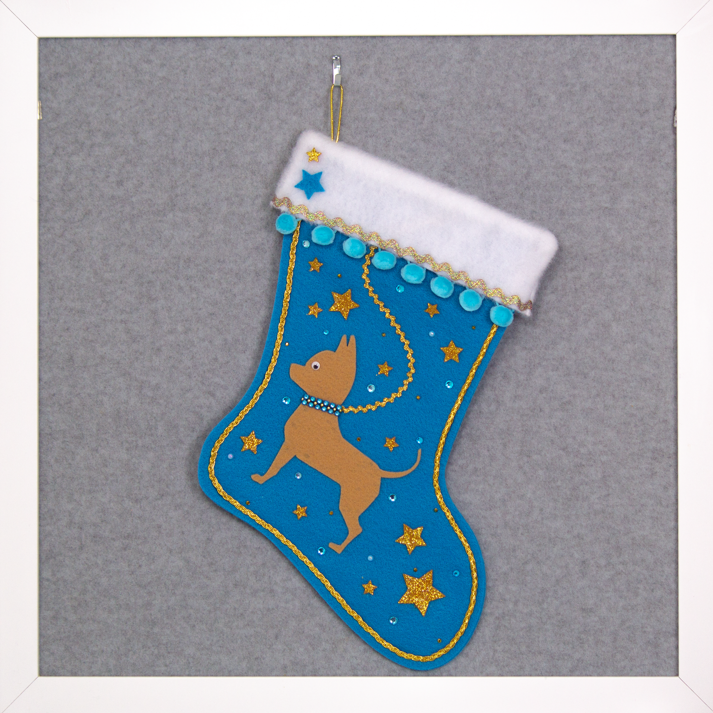 Tan short hair Chihuahua dog on peacock blue felt Christmas stocking with pom poms, glitter stars and decorations in blue and gold. (front of stocking)