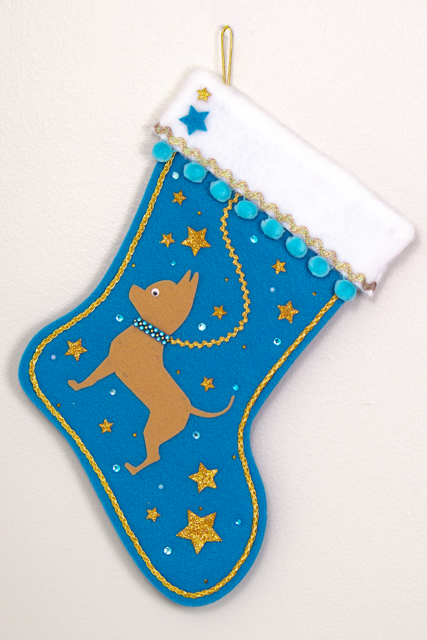 Tan short hair Chihuahua dog on peacock blue felt Christmas stocking with pom poms, glitter stars and decorations in blue and gold. (front of stocking)