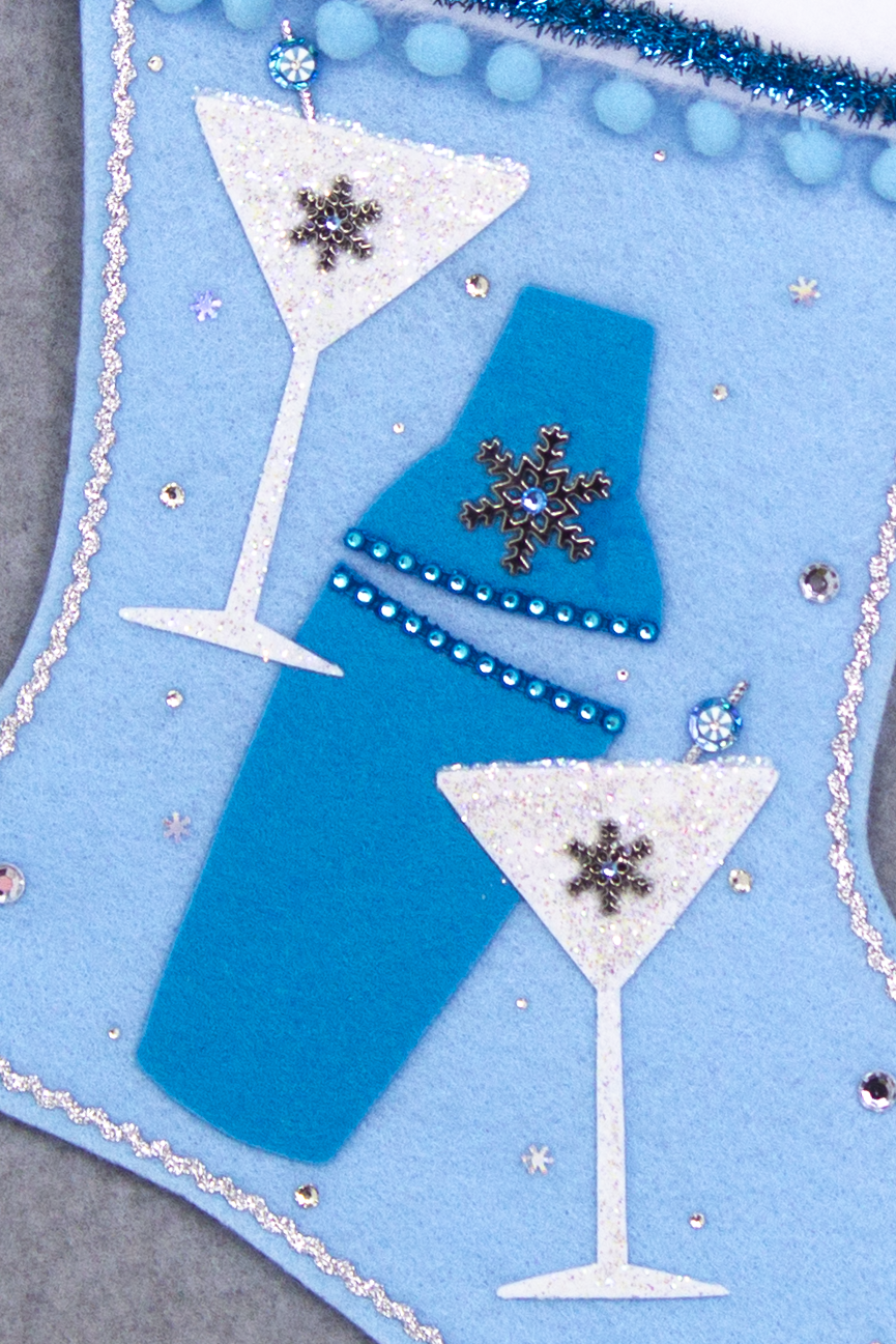 Blue felt shaker, chunky glitter painted white felt martini glasses on light blue felt handmade Christmas stocking with snowflake buttons, pom poms, tinsel ribbon and decorations in blues, white, clear & silver. (front of Cocktail stocking - close up detail of glasses and shaker)