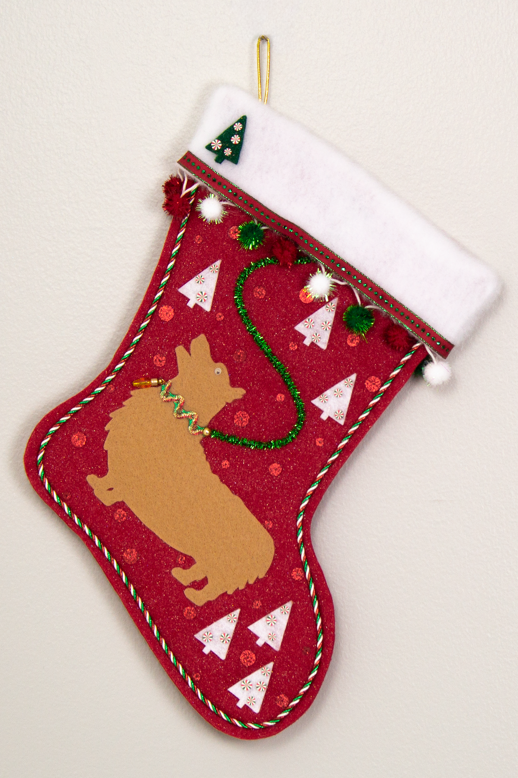 Tan Corgi on glitter red felt Christmas stocking with trees, pom poms and decorated in white, green, red and gold. (front of stocking)