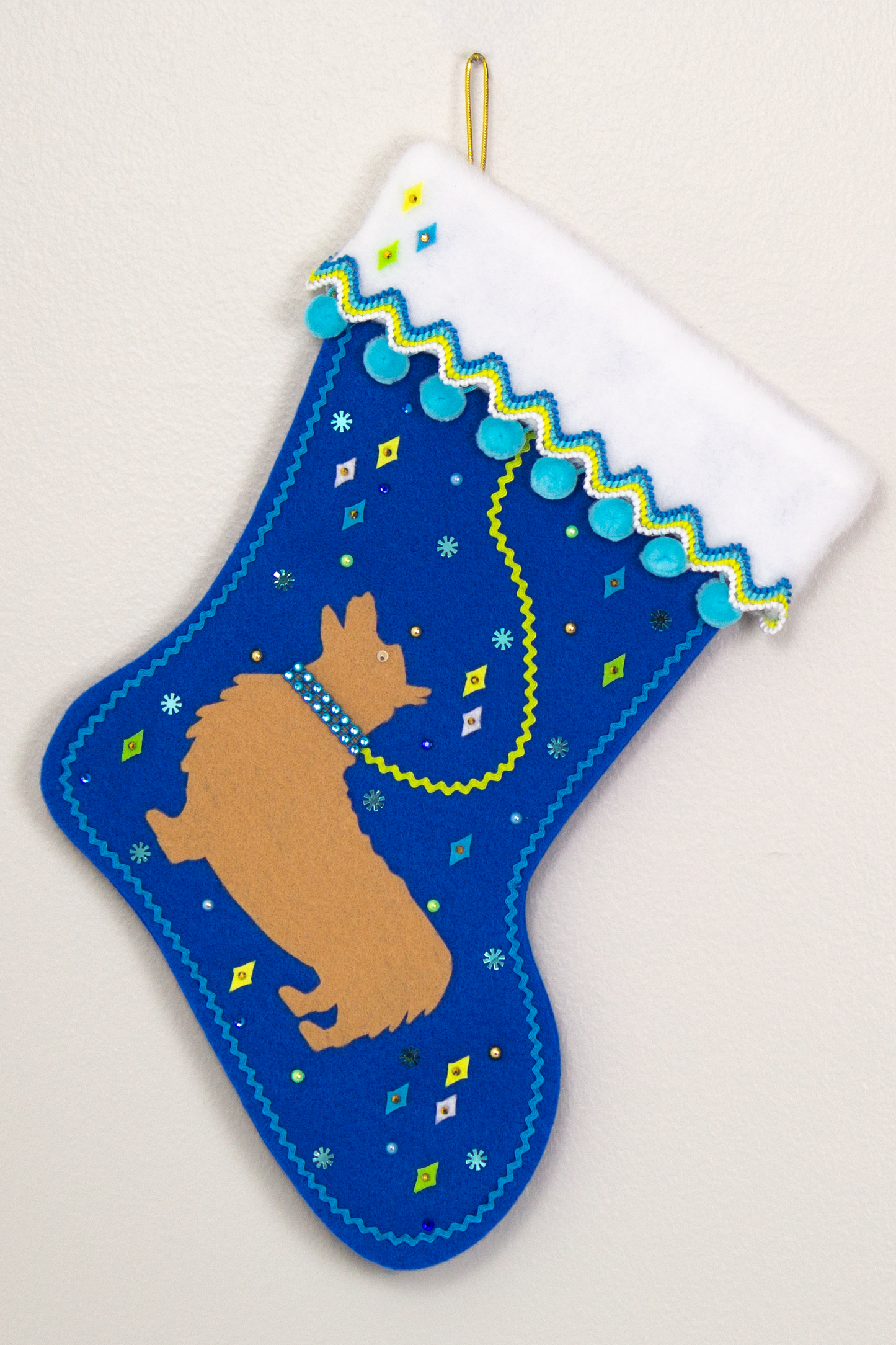 Tan Corgi dog on a royal blue felt Christmas stocking with pom poms, diamonds, snowflakes and decorated in yellow, green, blues and gold. (front of stocking)