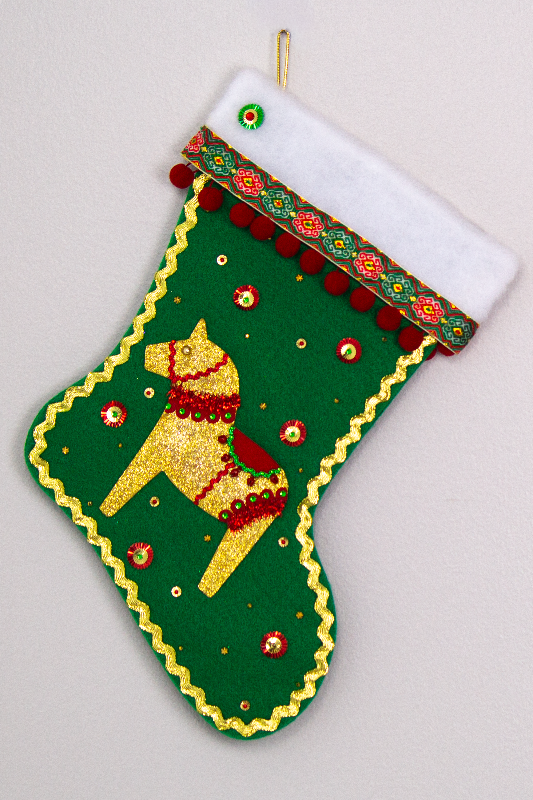 Gold glittered Dala horse on holiday green felt handmade Christmas stocking with pom poms, decorative ribbon, sunburst sequins and decorated in gold, red & green. (front of stocking)