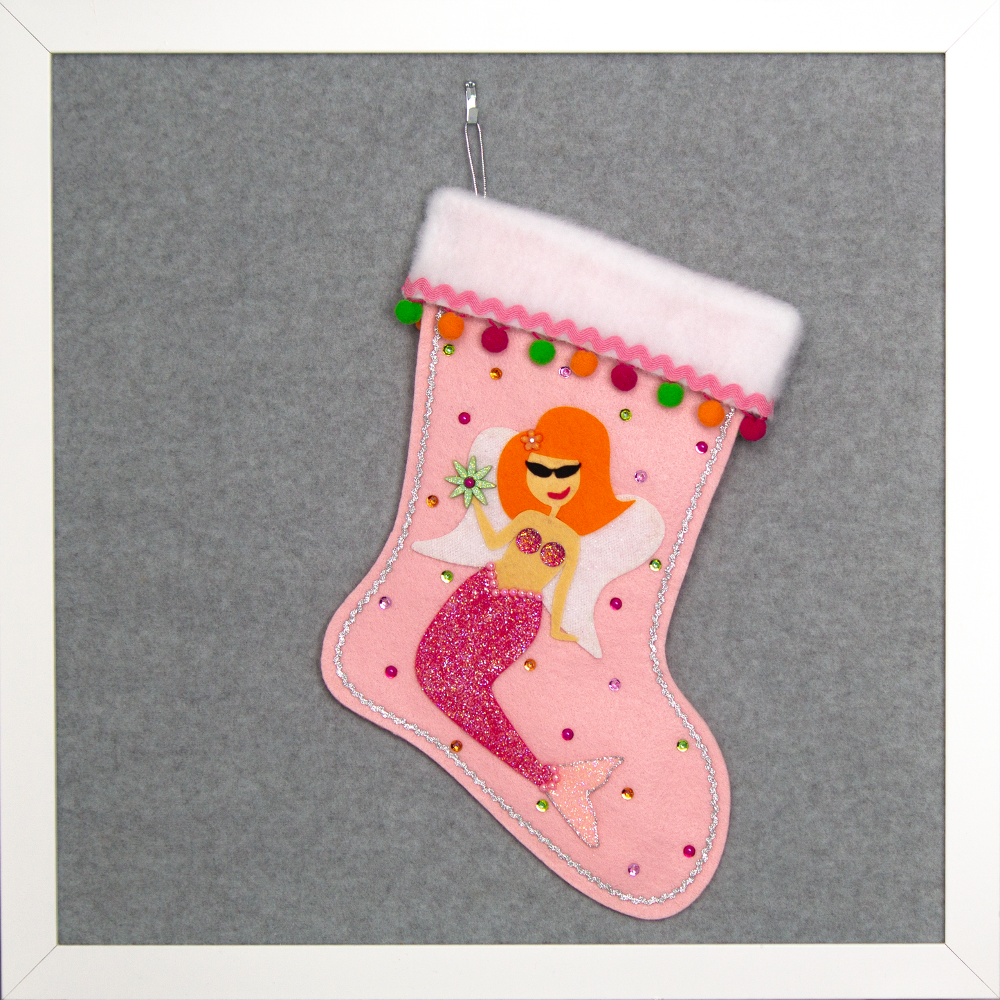 Ethel Mermaid with orange hair, glittered pink felt fin/tail, wings in white felt & glittery silver mesh, holding a sparkly flower button and a "bra" of  large multifaceted rhinestones on a light pink felt handmade Christmas stocking decorated in green, orange, pinks & silver. (front of stocking)