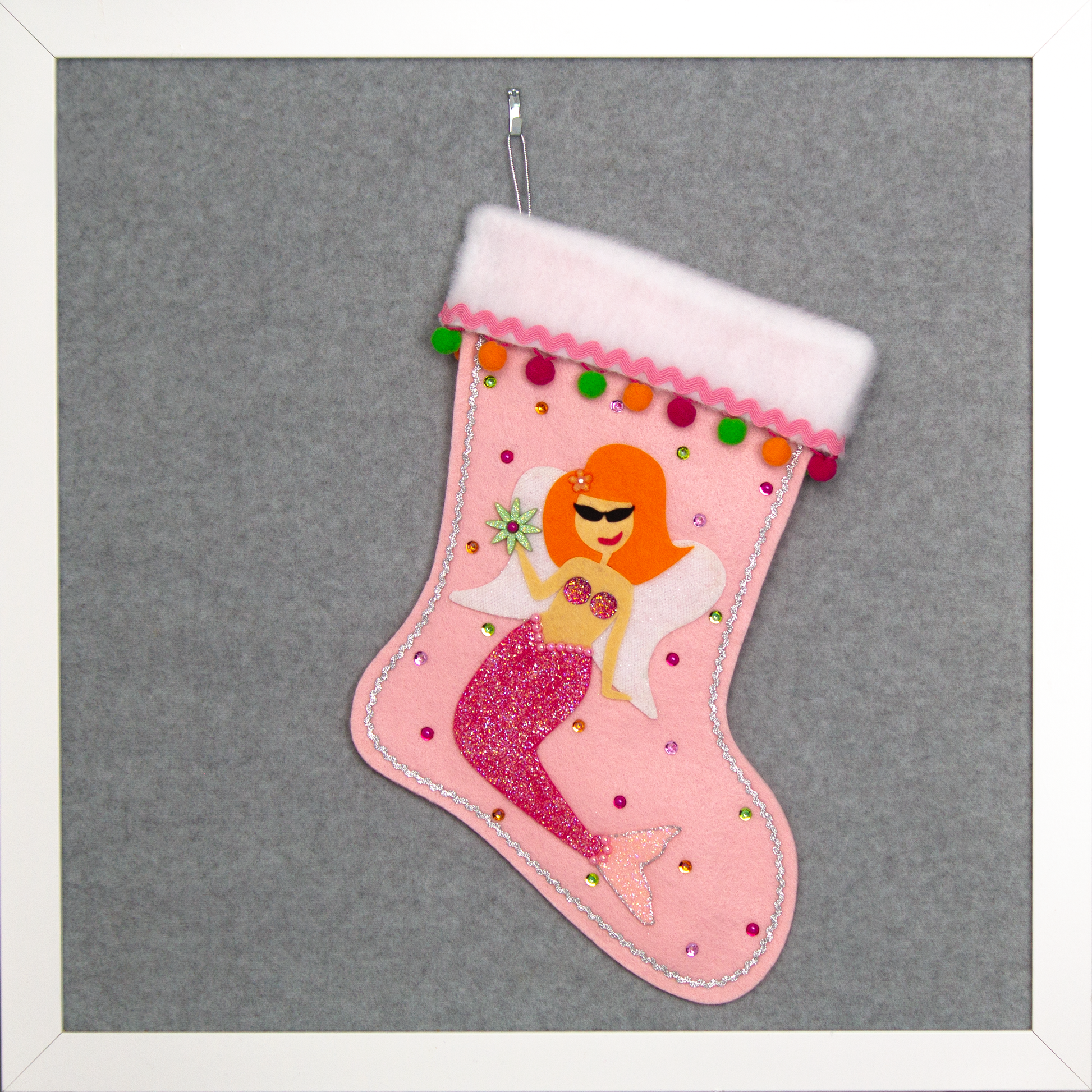 Ethel Mermaid with orange hair, glittered pink felt fin/tail, wings in white felt & glittery silver mesh, holding a sparkly flower button and a "bra" of  large multifaceted rhinestones on a light pink felt handmade Christmas stocking decorated in green, orange, pinks & silver. (front of stocking)