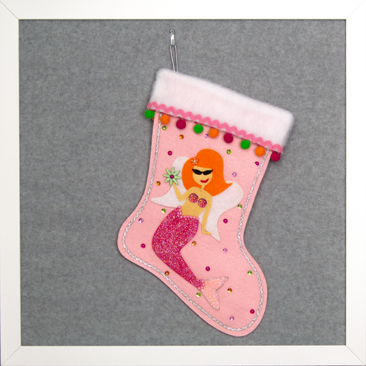 Ethel Mermaid with orange hair, glittered pink felt fin/tail, wings in white felt & glittery silver mesh, holding a sparkly flower button and a "bra" of  large multifaceted rhinestones on a light pink felt handmade Christmas stocking decorated in green, orange, pinks & silver. (front of stocking)