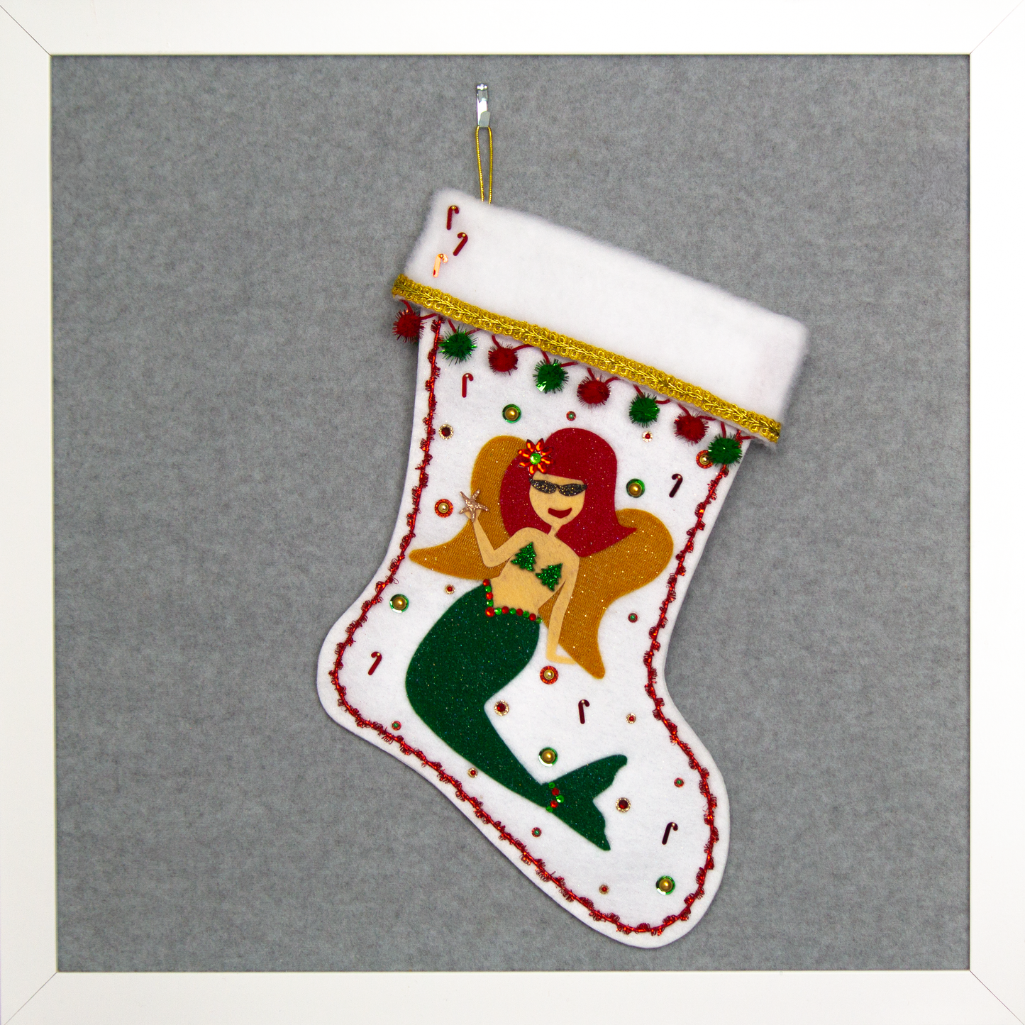 Ethel Mermaid with glitter felt red hair, dark green glitter felt fin/tail, wings in gold felt & glittery gold mesh, holding a sparkly starfish button and a "bra" of acrylic green glitter trees on a white glitter felt handmade Christmas stocking decorated in  red, green & gold. (front of stocking)