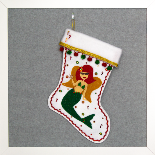 Ethel Mermaid with glitter felt red hair, dark green glitter felt fin/tail, wings in gold felt & glittery gold mesh, holding a sparkly starfish button and a "bra" of acrylic green glitter trees on a white glitter felt handmade Christmas stocking decorated in  red, green & gold. (front of stocking)