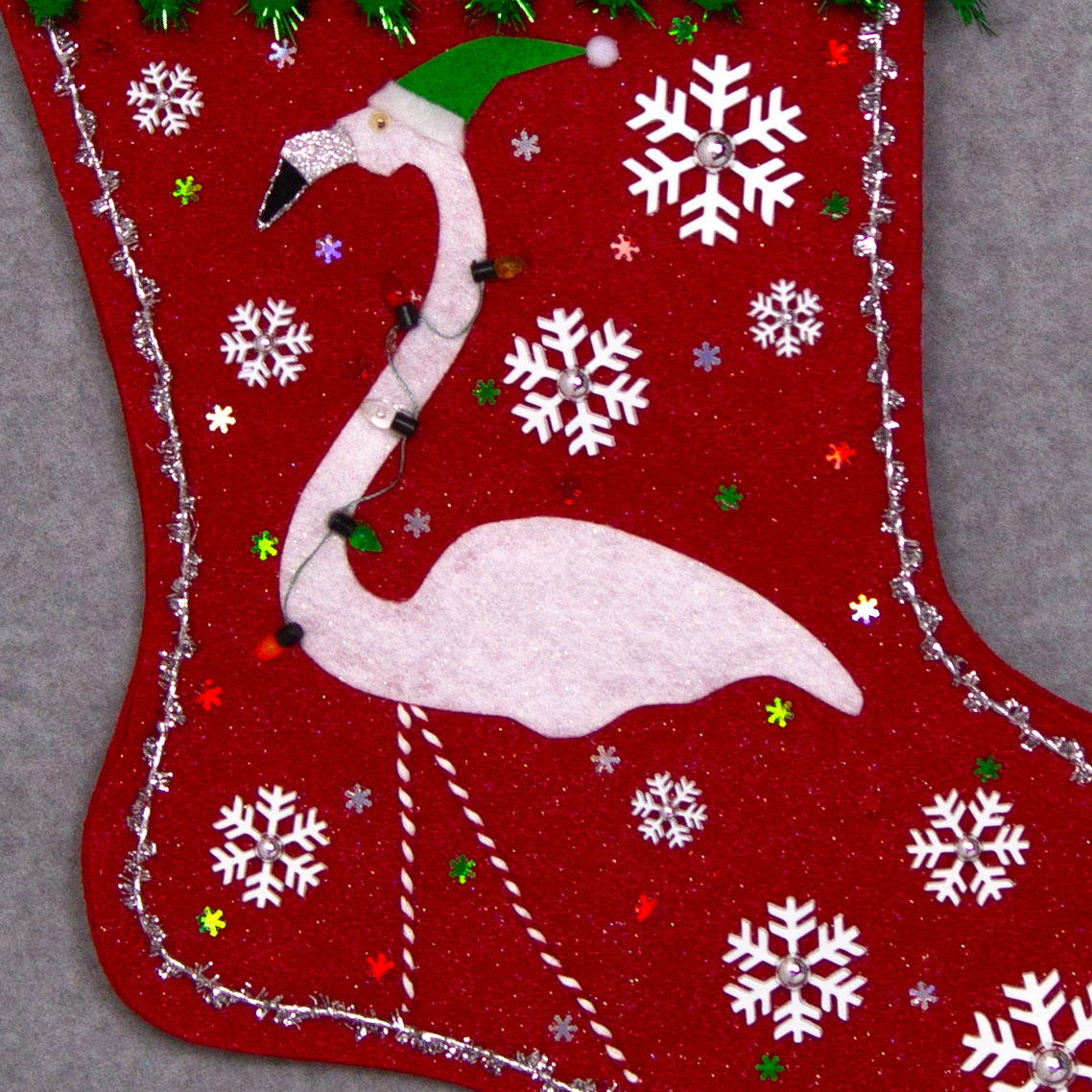White glitter felt flamingo on red glitter felt handmade Christmas stocking with sparkly pom poms, snowflake buttons, fake lights and decorated in white, green, red & silver. (front of stocking - close up detail of flamingo)