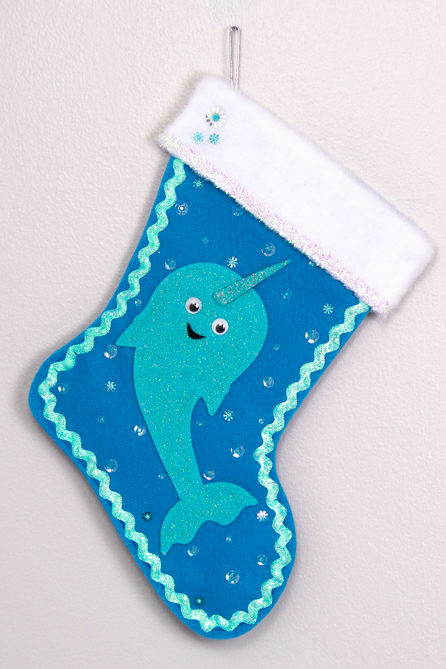 Glittery aqua Narwhal on a peacock blue felt handmade Christmas stocking with tinsel ribbon, googlie eyes and decoration in clear, aqua & silver. (front of stocking)