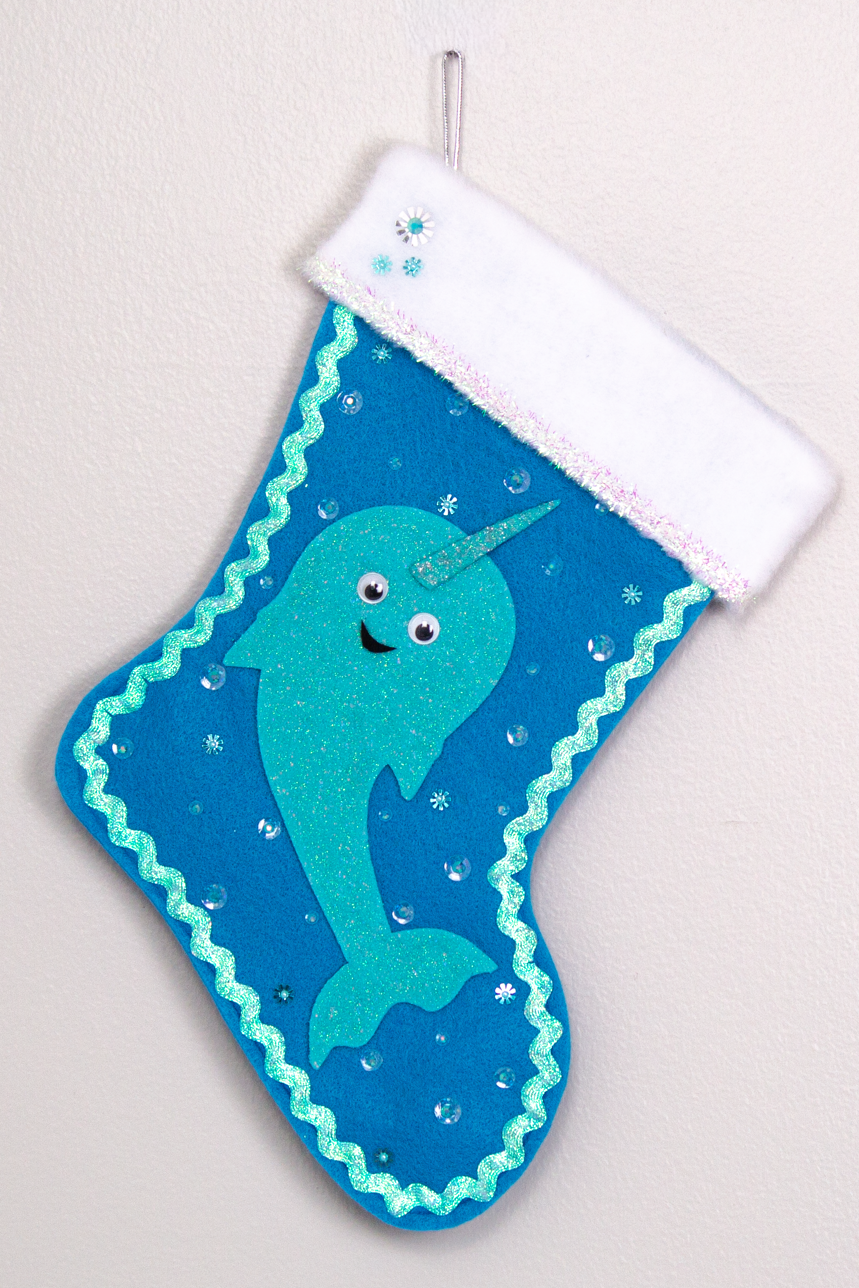 Glittery aqua Narwhal on a peacock blue felt handmade Christmas stocking with tinsel ribbon, googlie eyes and decoration in clear, aqua & silver. (front of stocking)