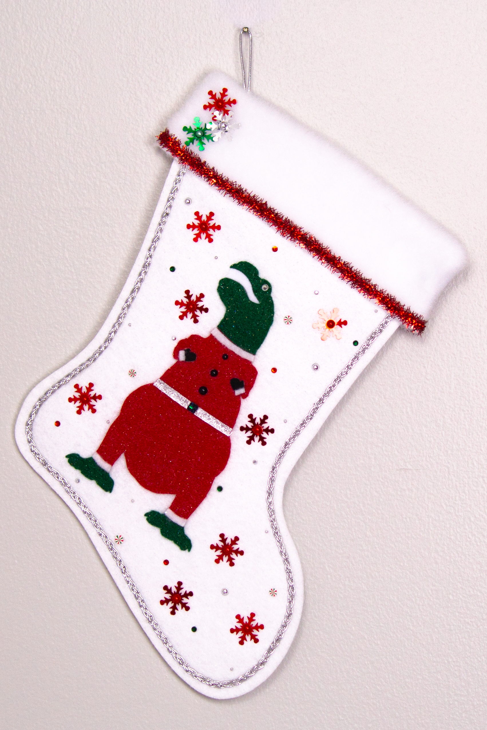 Dark green glitter felt T-Rex with red glitter suit on white glitter felt handmade Christmas stocking with tinsel ribbon, snowflake sequins and decoration in red, green & silver. (front of stocking)