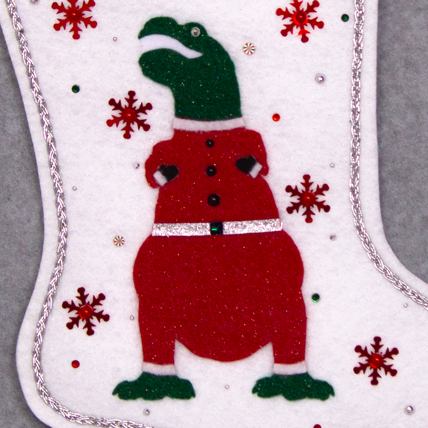 Dark green glitter felt T-Rex with red glitter suit on white glitter felt handmade Christmas stocking with tinsel ribbon, snowflake sequins and decoration in red, green & silver. (front of stocking - close up detail of T-Rex)