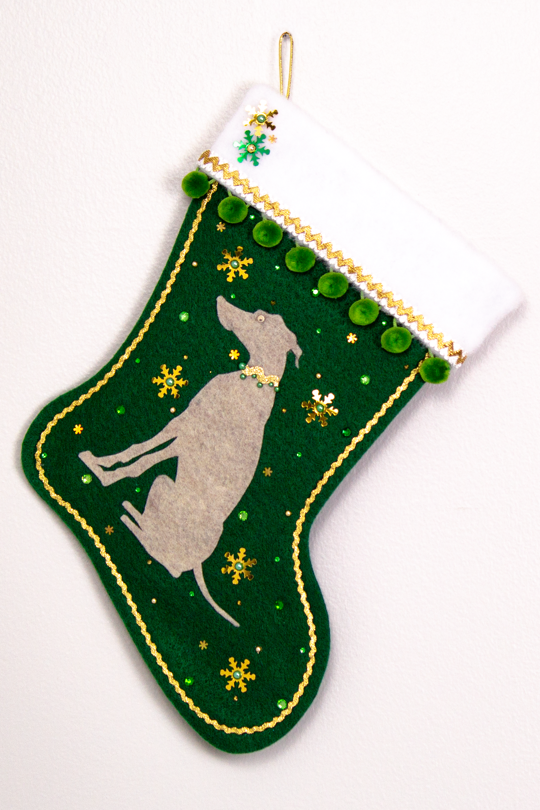 Sandstone beige Whippet dog on dark green felt handmade Christmas stocking with snowflake sequins, pom poms and decorated in gold and green. (front of stocking)